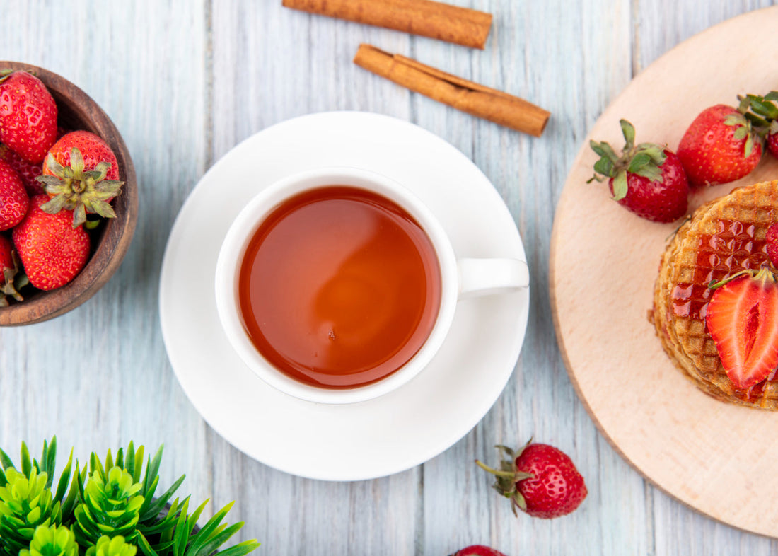 Cup of tea on wooden table with fresh strawberries, waffles topped with strawberry syrup, and cinnamon sticks