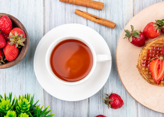 Cup of tea on wooden table with fresh strawberries, waffles topped with strawberry syrup, and cinnamon sticks