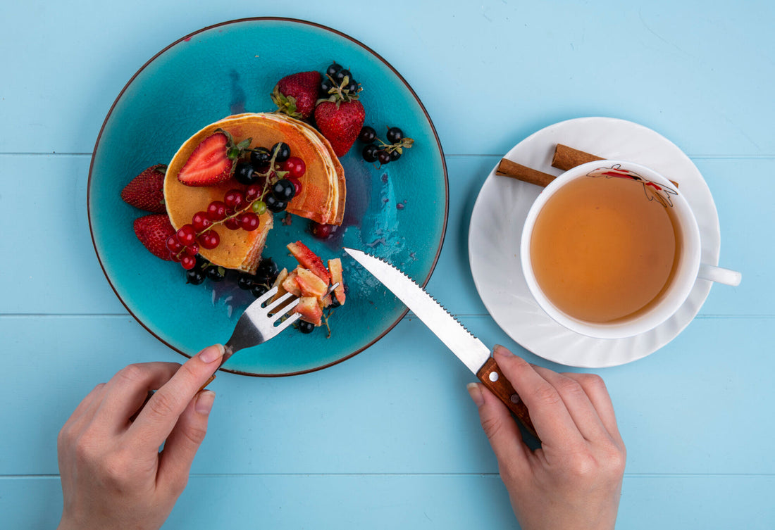 Person cutting pancakes with mixed berries on blue plate and cup of tea with cinnamon sticks