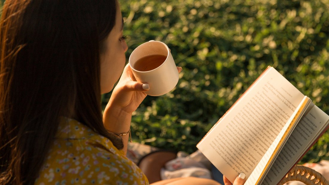Woman in yellow floral dress reading book and holding coffee cup outdoors on grass