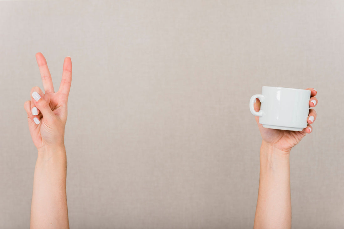 Two hands against beige background, left showing peace sign, right holding white coffee mug