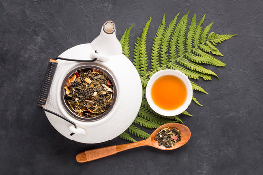 White teapot with loose leaf tea, wooden spoon with tea, cup of brewed tea on green fern over dark surface