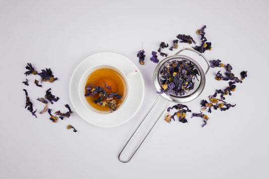 White cup of herbal tea with butterfly pea flowers and tea strainer on white background