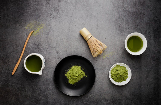 Matcha green tea powder on black and white plates, bamboo whisk, and tea in cups on dark textured surface