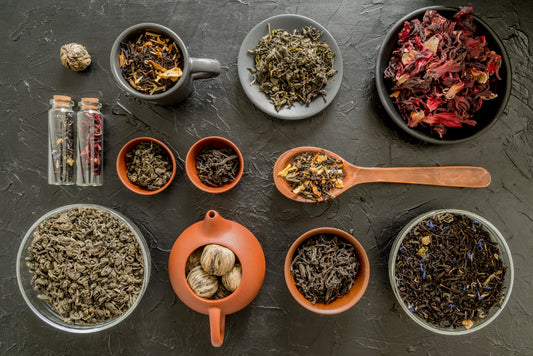 Various loose leaf teas, dried flowers, and tea balls displayed in bowls, bottles, and a wooden spoon on a textured dark surface