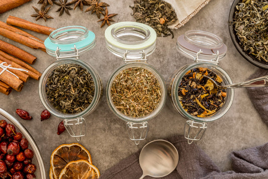 Three glass jars with assorted loose leaf herbal and black tea blends surrounded by spices and dried citrus on gray surface