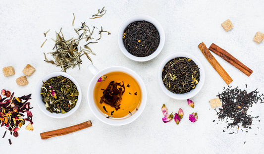 Flat lay of tea leaves in bowls, brewed tea cup, cinnamon sticks, sugar cubes, and rosebuds on white surface