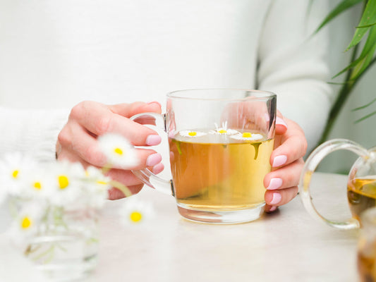 Person holding clear glass cup of chamomile tea with floating flowers on white table