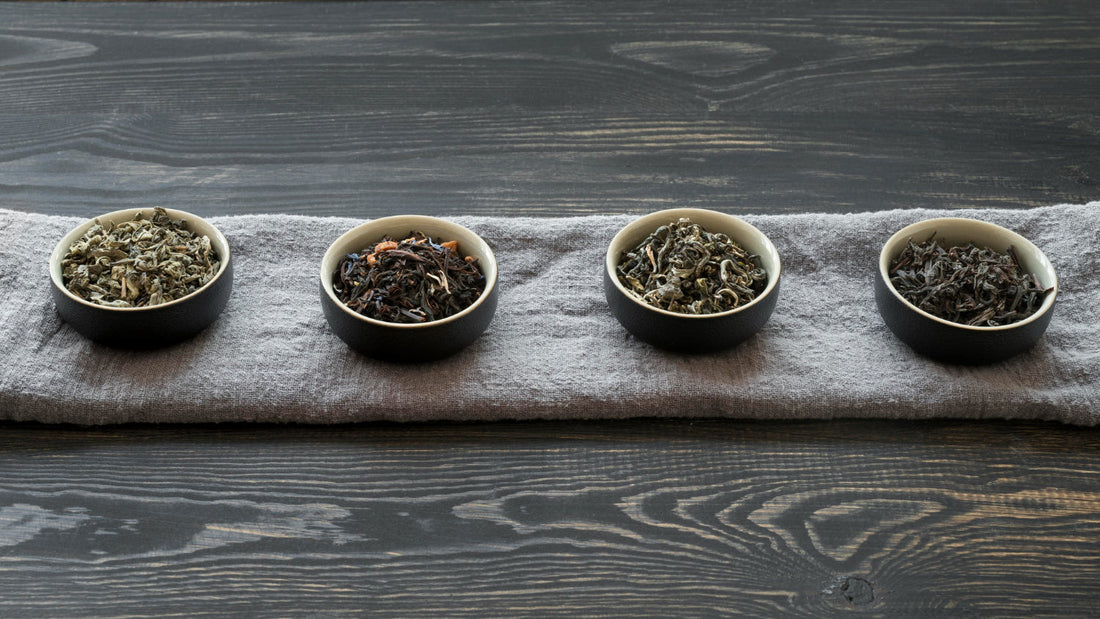 Four black bowls with different types of loose leaf tea on gray cloth over dark wooden table