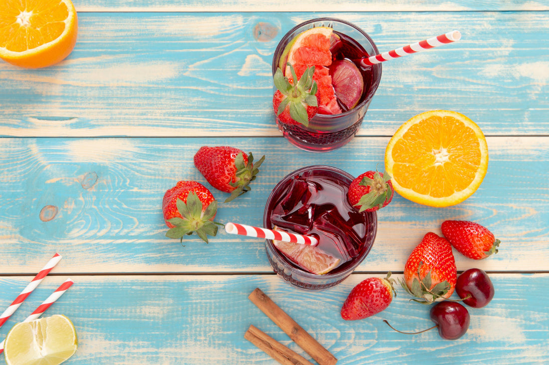 Two glasses of red fruit punch with ice and striped straws on blue wooden table surrounded by strawberries, orange halves, cherries, and cinnamon sticks