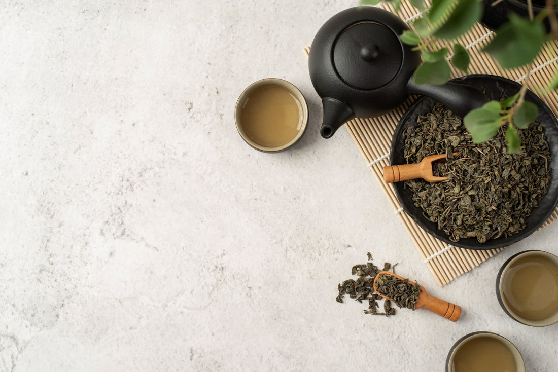 Black teapot with three cups of brewed green tea and loose green tea leaves on a light textured surface
