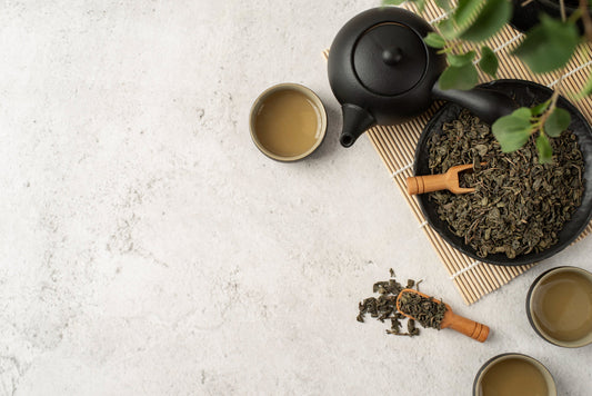 Black teapot with three cups of brewed green tea and loose green tea leaves on a light textured surface
