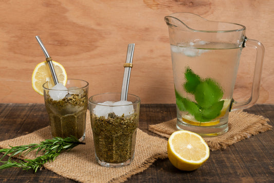 Two glasses of iced yerba mate with metal straws, lemon slices, pitcher with mint and lemon, rosemary on wooden table