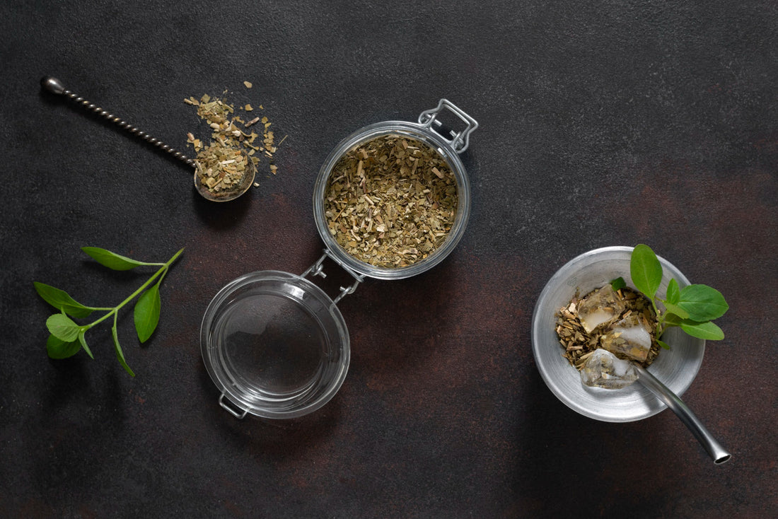 Yerba mate leaves in glass jar and metal cup with ice and green sprigs on dark surface