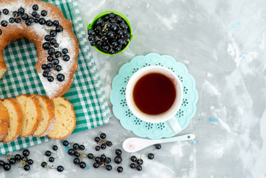 Sliced bundt cake with powdered sugar and blackcurrants on checkered cloth with cup of tea on blue doily