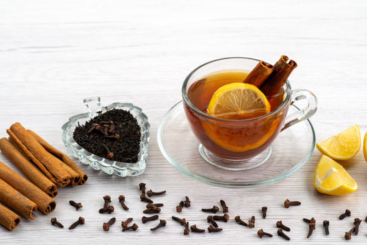 Glass cup of black tea with lemon slice and cinnamon sticks, surrounded by loose tea, cloves, cinnamon, and lemon wedges