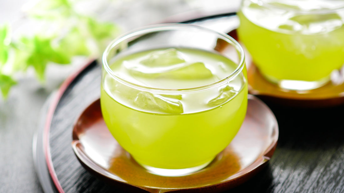 Glass cup of iced green tea on wooden saucer with blurred second cup and green leaves background