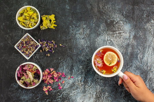 Hand holding a white cup of herbal tea with lemon slice and edible flowers on dark textured background with dried flower petals in white bowls