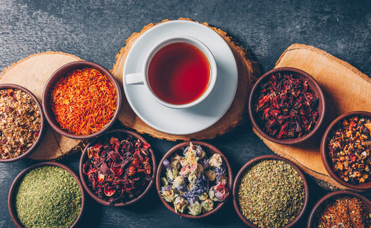White cup of herbal tea on wooden coaster surrounded by various dried herbs and spices in bowls