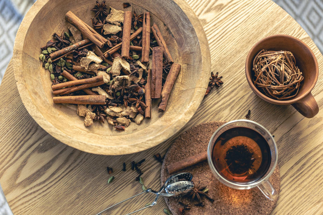Wooden bowl with cinnamon sticks, star anise, ginger, cardamom on wooden table with tea cup and infuser
