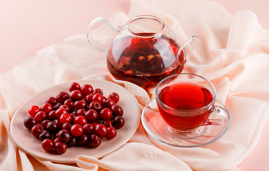 Clear glass teapot and cup with red berry tea next to plate of fresh cranberries on soft fabric