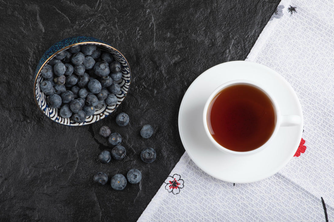 Cup of black tea on white saucer next to blue patterned bowl of fresh blueberries on dark surface
