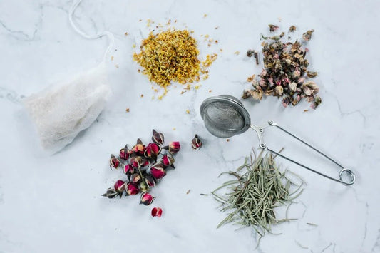 Herbal tea ingredients including dried rosebuds, yellow flowers, lavender, with tea infuser and tea bag on marble surface