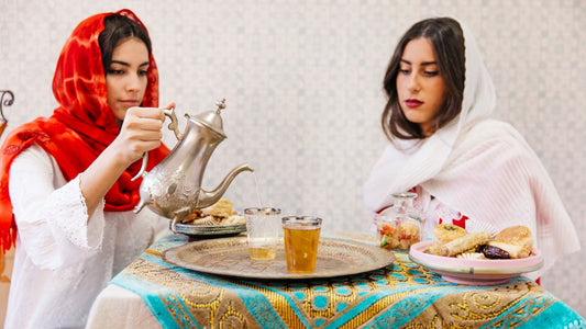Two women in traditional shawls pouring and enjoying Kashmiri Kahwa tea with snacks on a decorative table