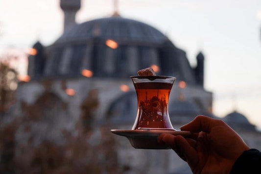 Hand holding traditional Turkish tea in glass with blurred mosque and lights in background at dusk