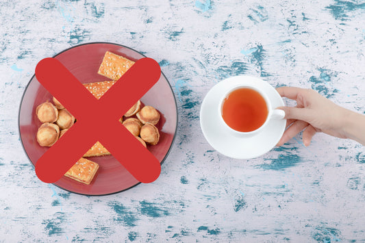 Hand holding white cup of tea on saucer near plate of cookies crossed out with red X on blue rustic table
