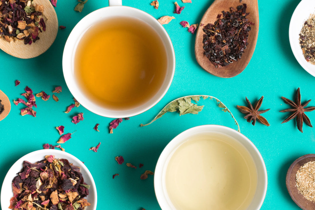 Two cups of herbal tea surrounded by dried tea leaves, spices, and flower petals on teal background