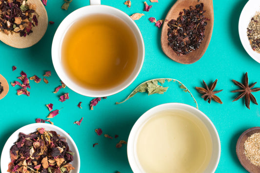 Two cups of herbal tea surrounded by dried tea leaves, spices, and flower petals on teal background