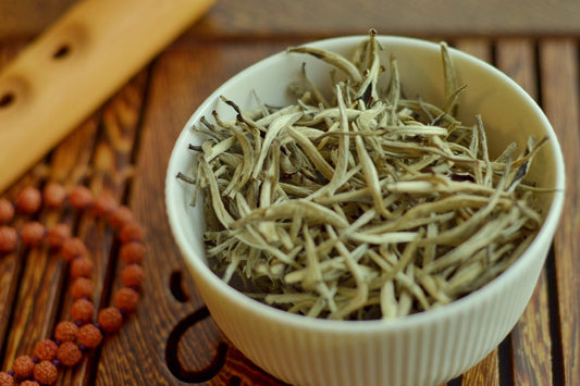 Loose white tea leaves in a white bowl on a wooden surface with a wooden flute and a brown beaded bracelet